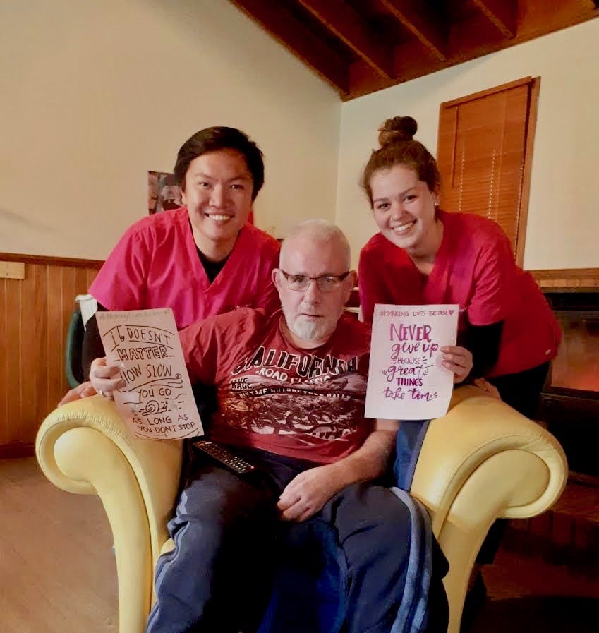Nurse Next Door client Mick sitting in his armchair at home with two of his 24/7 caregiving team helping him write signs of affirmation and possibility to maintain his positive outlook on life